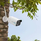 KAAMERAMAAILM.EE - Valge Valvekaamera Reolink RLC-511WA 5MP Dual Band WiFi, 5X Zoom on paigaldatud õues kiviseinale, mida raamivad rohelised lehed ja selge sinine taevas taustal. (5/5)