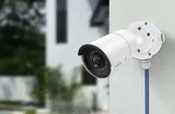 KAAMERAMAAILM.EE - Valge Reolink B10 Ühenduskarp torukaameratele on paigaldatud välisseinale, suunaga väljapoole. Hämarad moodsad hooned ja rohelised lehed taustal rõhutavad turvalist ja stiilset kesk (3/3)